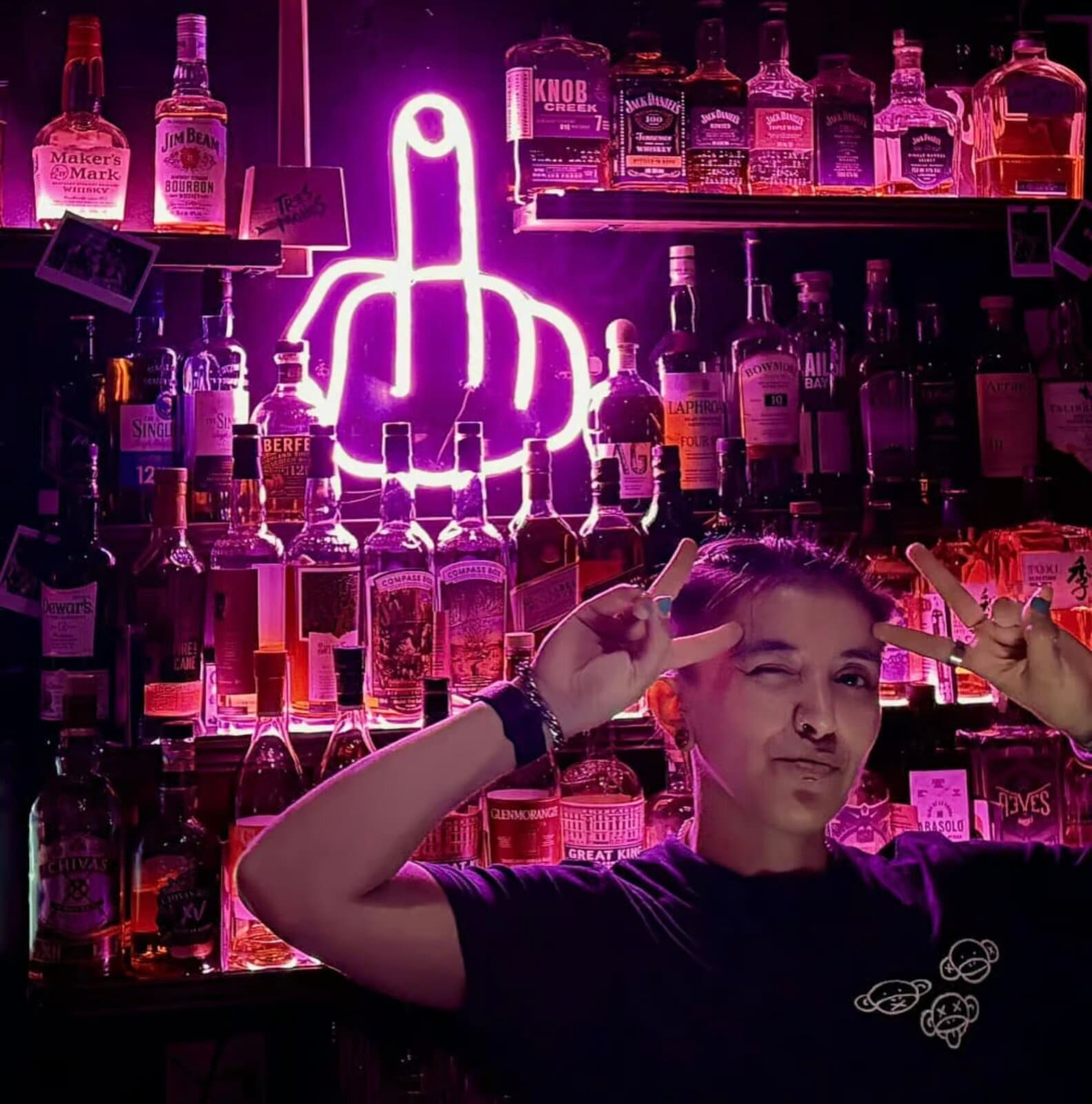 A bartender at Tres Monos throwing devil horns beneath a neon middle-finger sign and shelves of whisky.