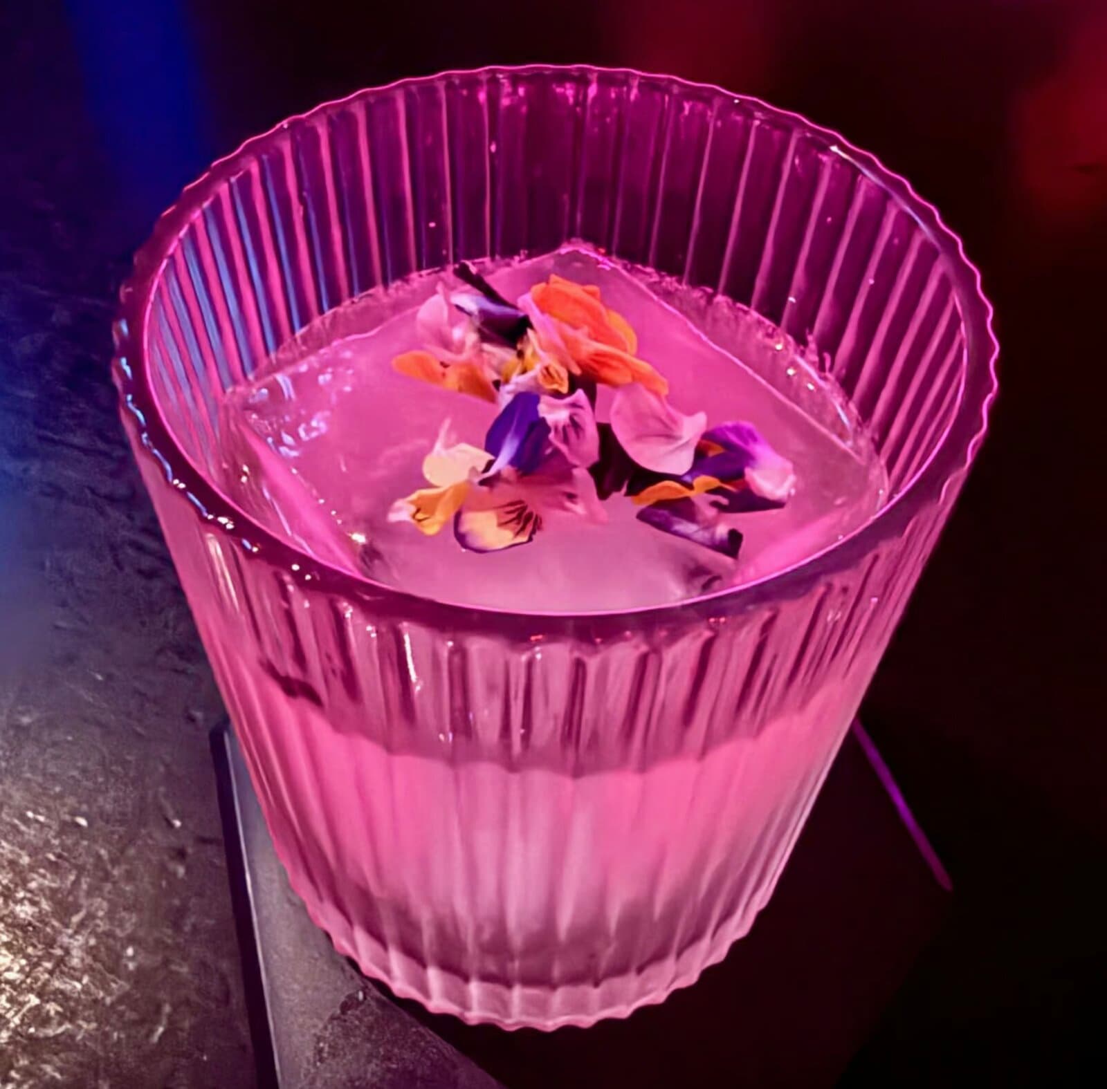 A pink Tres Monos cocktail in a fluted glass topped with edible flowers, under neon light.