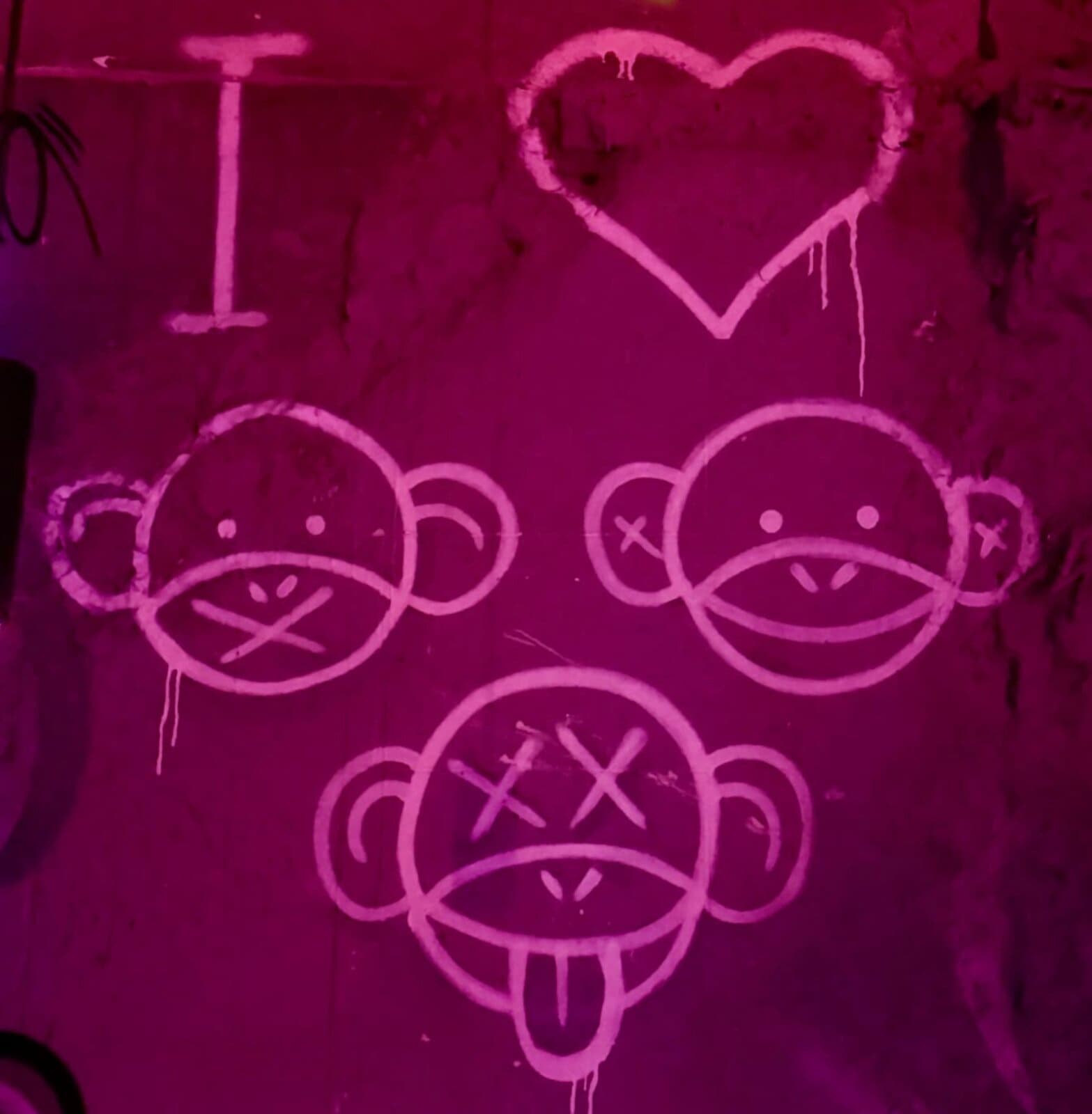 Tres Monos wall art: an I and a heart above three hand-drawn monkey faces in pink UV paint.